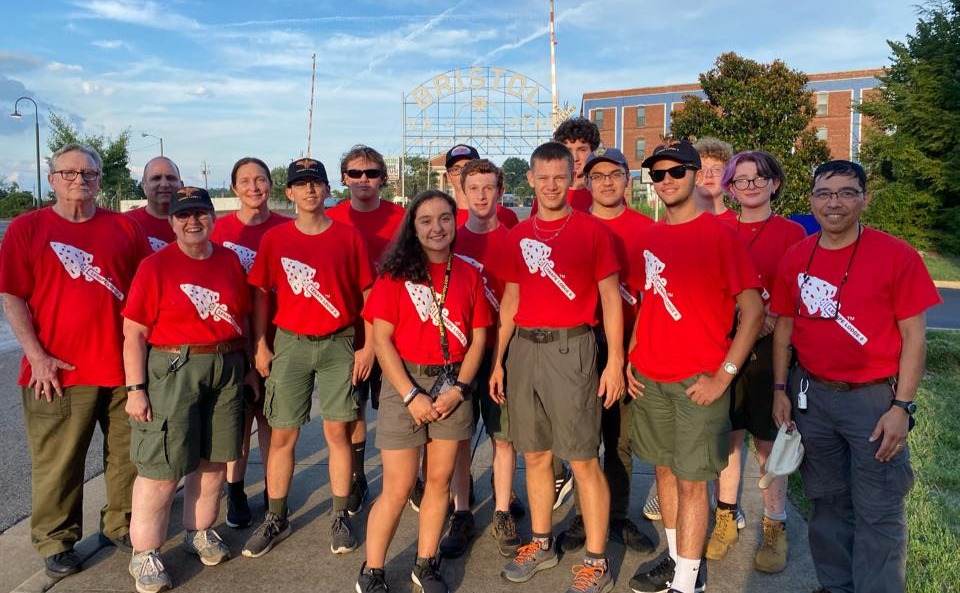 Teenaged Scouts and adult volunteers wearing matching red shirts smile at the camera while standing outside on a sunny day.